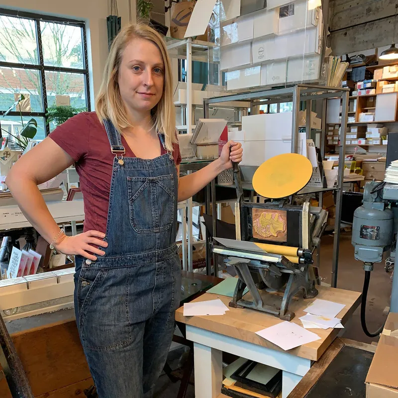 The artist standing next to a tabletop letterpress in a print studio