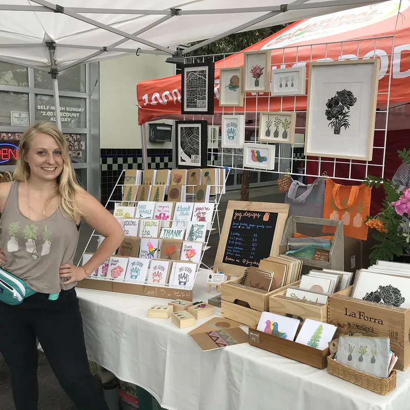 The artist at a farmers market booth with framed prints and racks of greeting cards