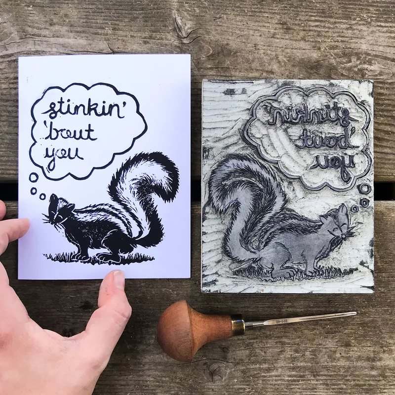 Skunk block print card reading “stinkin’ ’bout you” beside its carved printing block