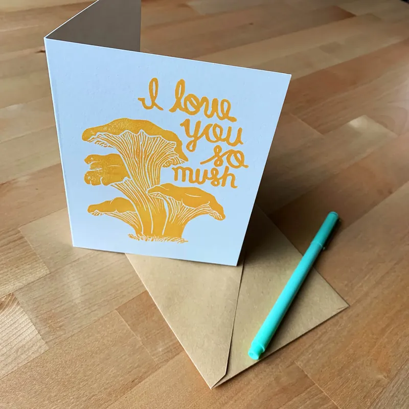 Folded card with a golden chanterelle mushroom and “I love you so much” lettering, with a teal pen on a kraft envelope